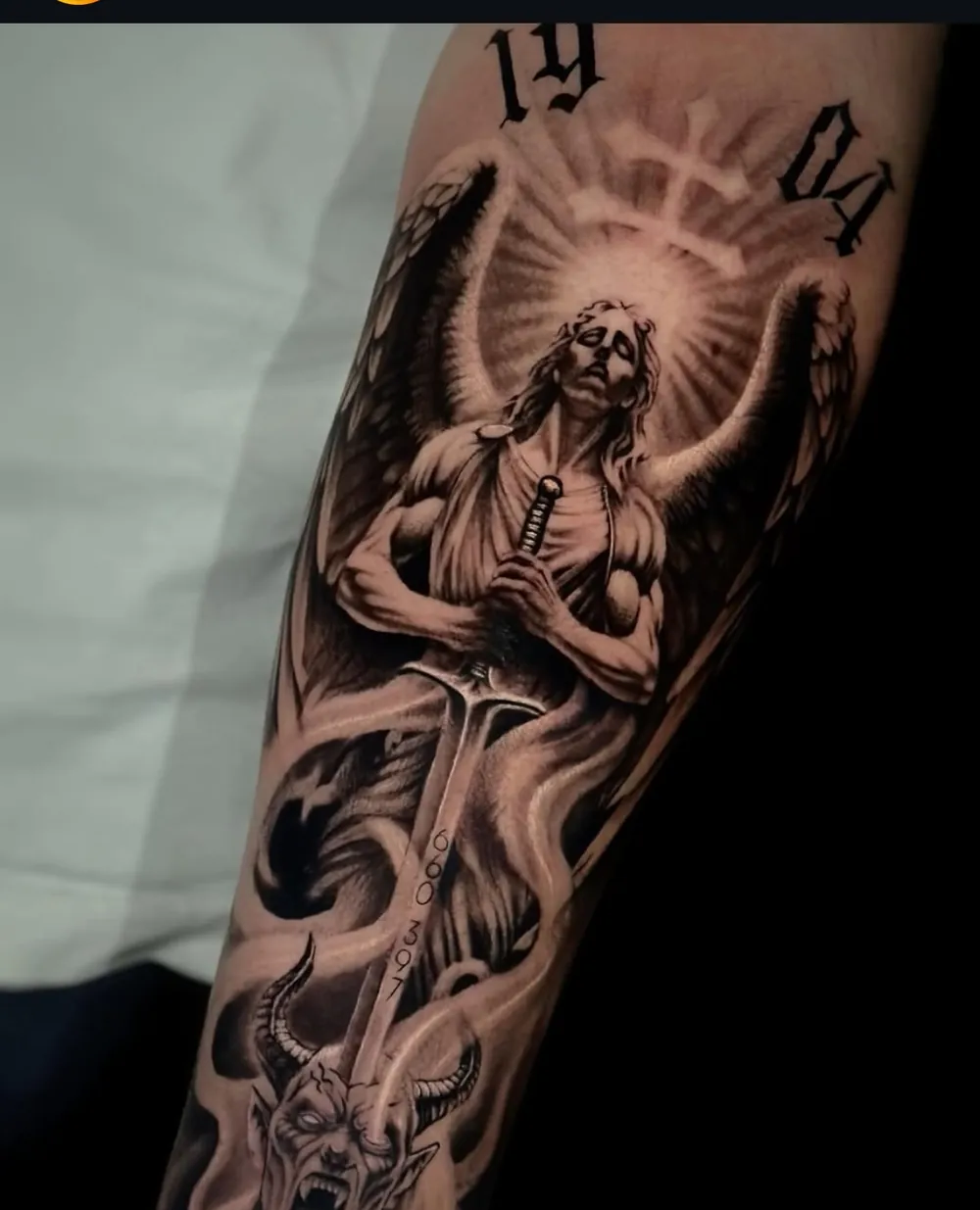 Antebrazo tatuado con un ángel con espada sobre un demonio, en negro y gris