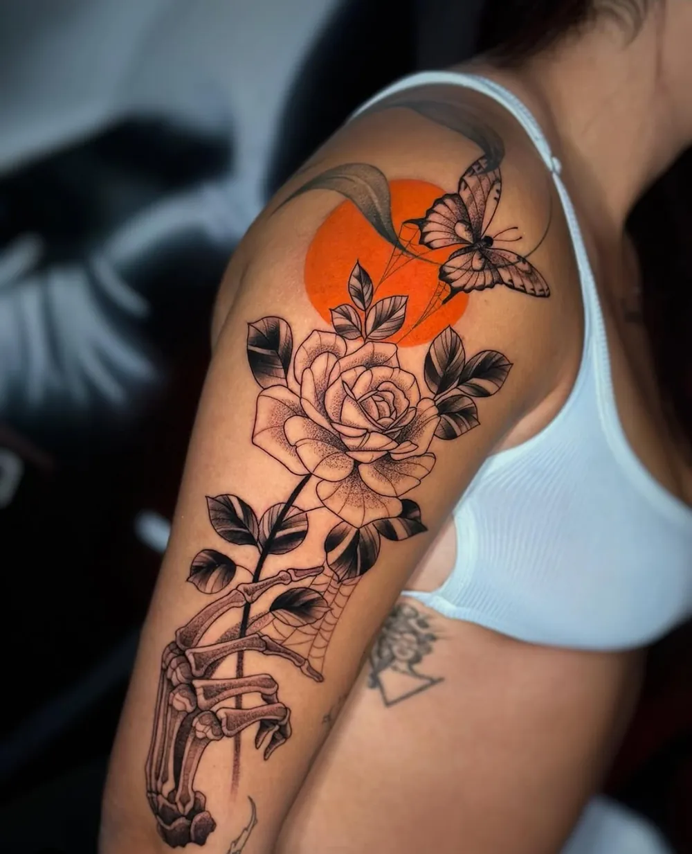 Tatuaje de una rosa en fine line y dotwork con una mano esquelética, una mariposa y un sol naranja